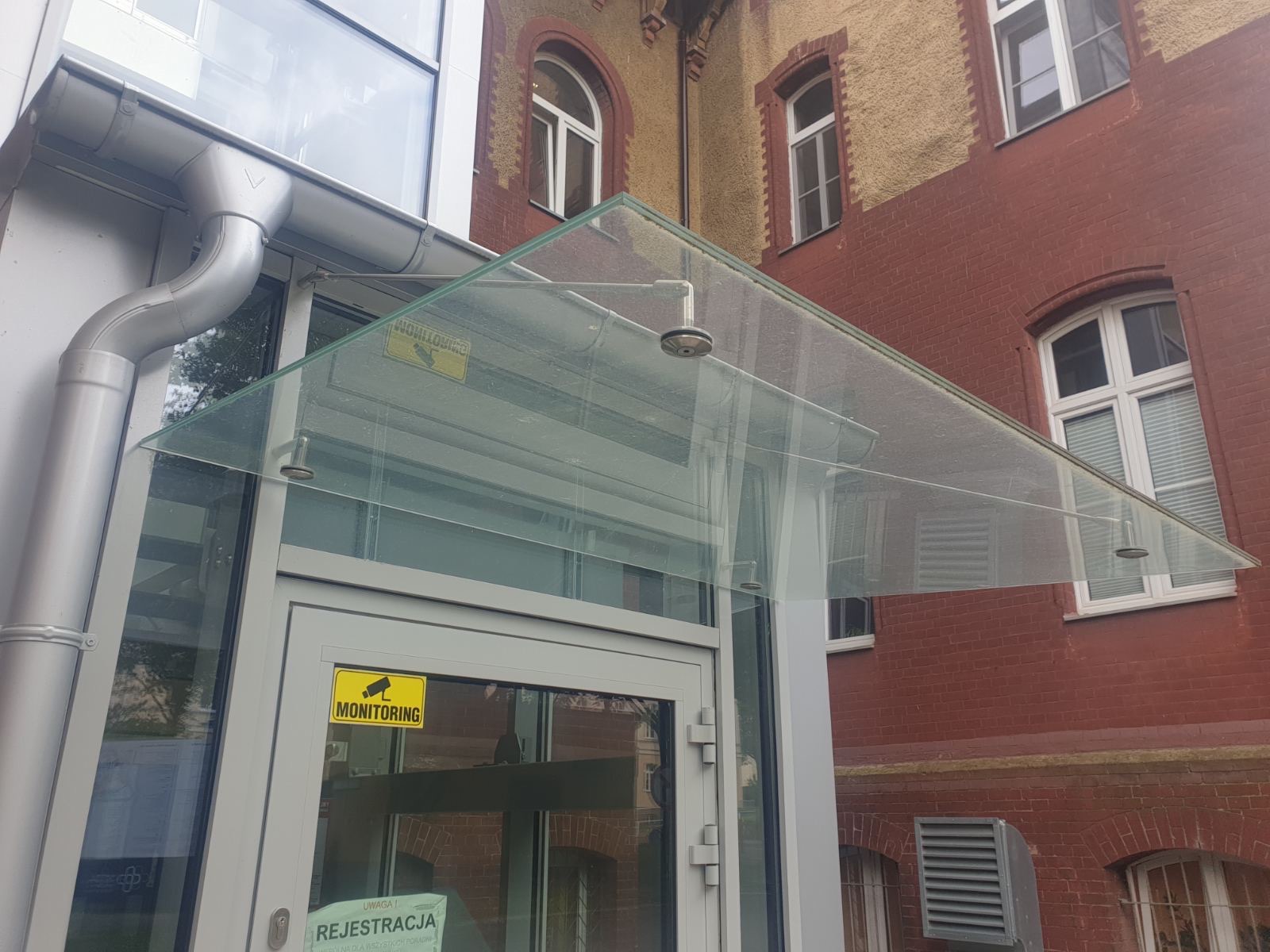 Szklany daszek nad wejściem do budynku, wsparty na metalowych elementach mocujących, z widocznym systemem rynnowym i tabliczką informacyjną o monitoringu.