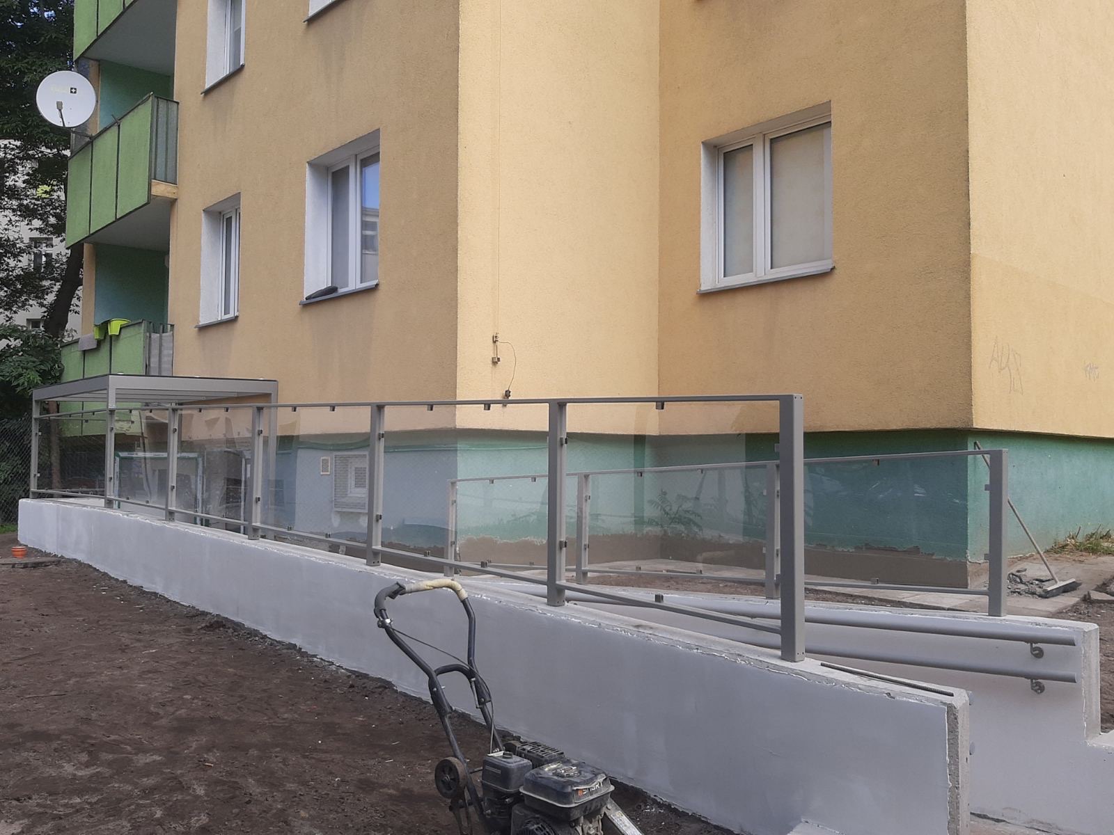 Szklana balustrada zamontowana na betonowym podwyższeniu przed budynkiem mieszkalnym, z widoczną kosiarką w pierwszym planie.