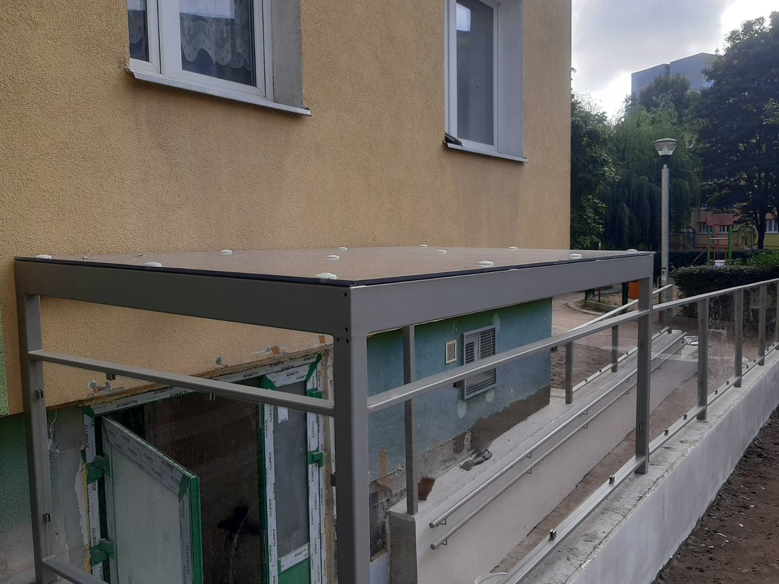 Zadaszenie nad zejściem do piwnicy z balustradą szklaną, widok z boku, elementy montażowe widoczne, budynek otynkowany w tle.
