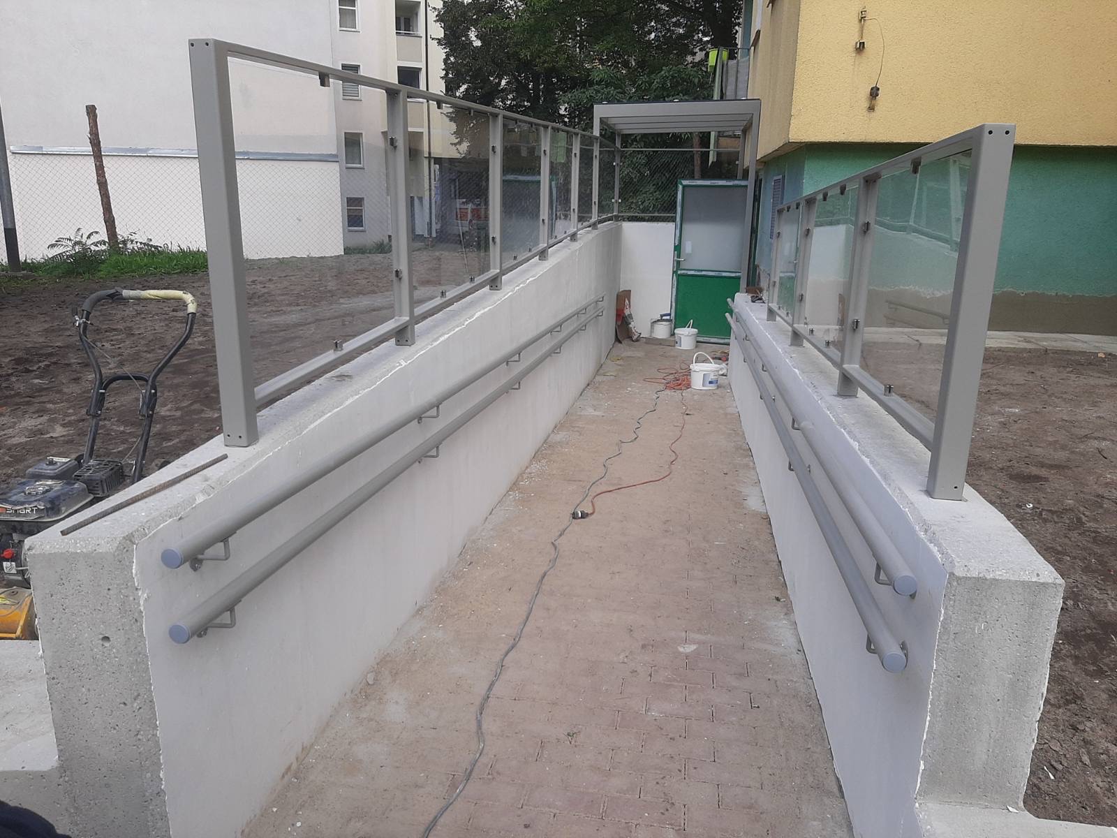Zewnętrzna rampa dla niepełnosprawnych z szarą, metalową balustradą szklaną i podwójną poręczą, widoczne ślady prac budowlanych, w tle budynek mieszkalny.