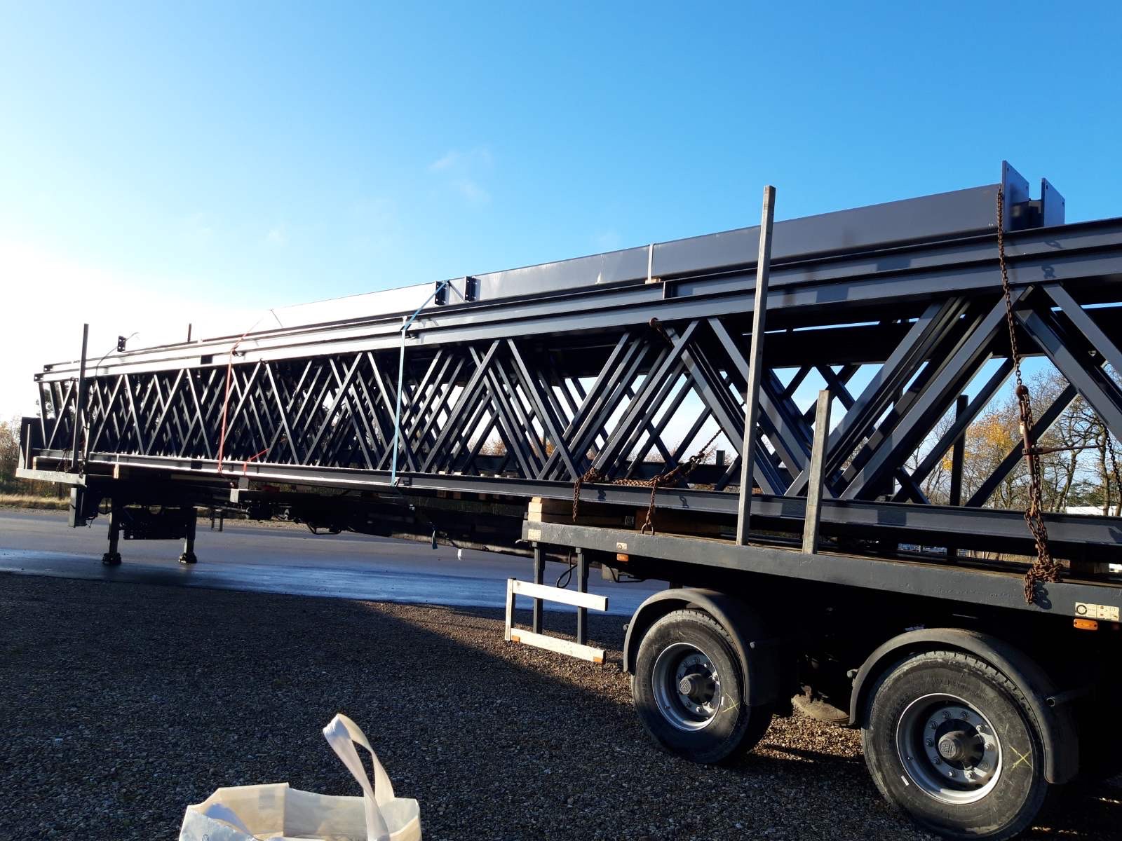 Gotowa, pomalowana na czarno konstrukcja stalowa w kształcie kratownicy, zabezpieczona łańcuchami na naczepie ciężarówki, gotowa do transportu, na tle błękitnego nieba.
