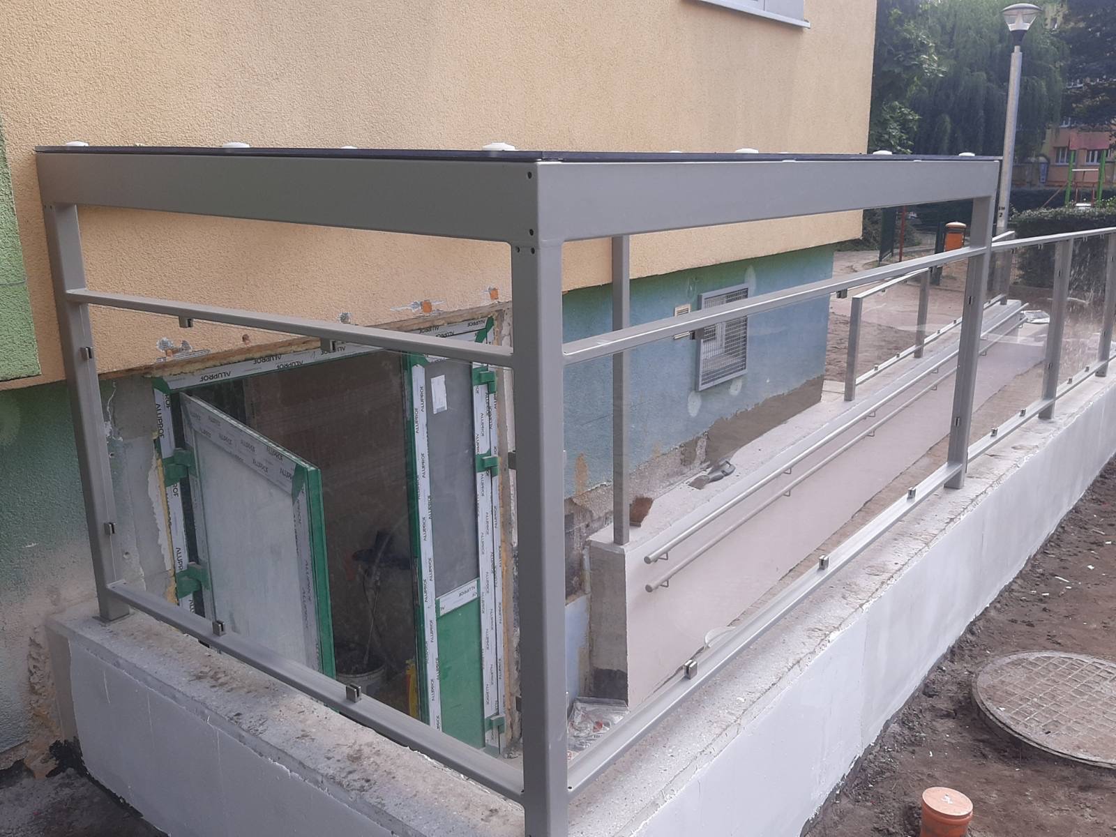 Szara metalowa konstrukcja z przeszklonymi panelami tworząca zadaszenie i balustradę nad wejściem do piwnicy budynku z widocznymi śladami prac budowlanych.