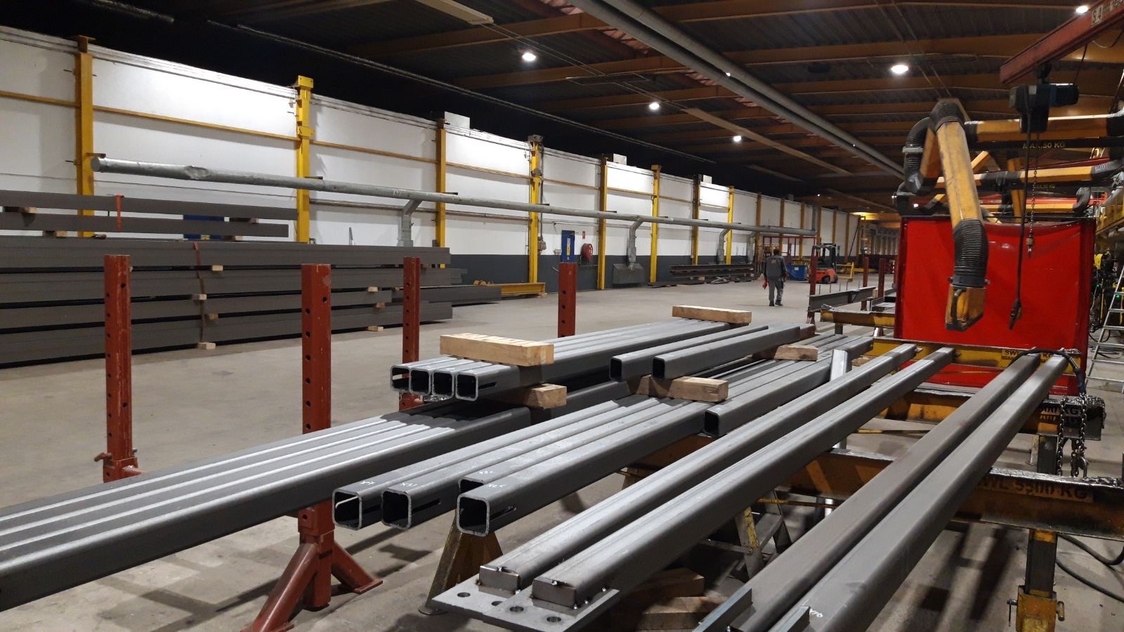 Stalowe profile kwadratowe, ułożone na podporach w hali produkcyjnej, w tle widoczny wózek widłowy i pracownik.