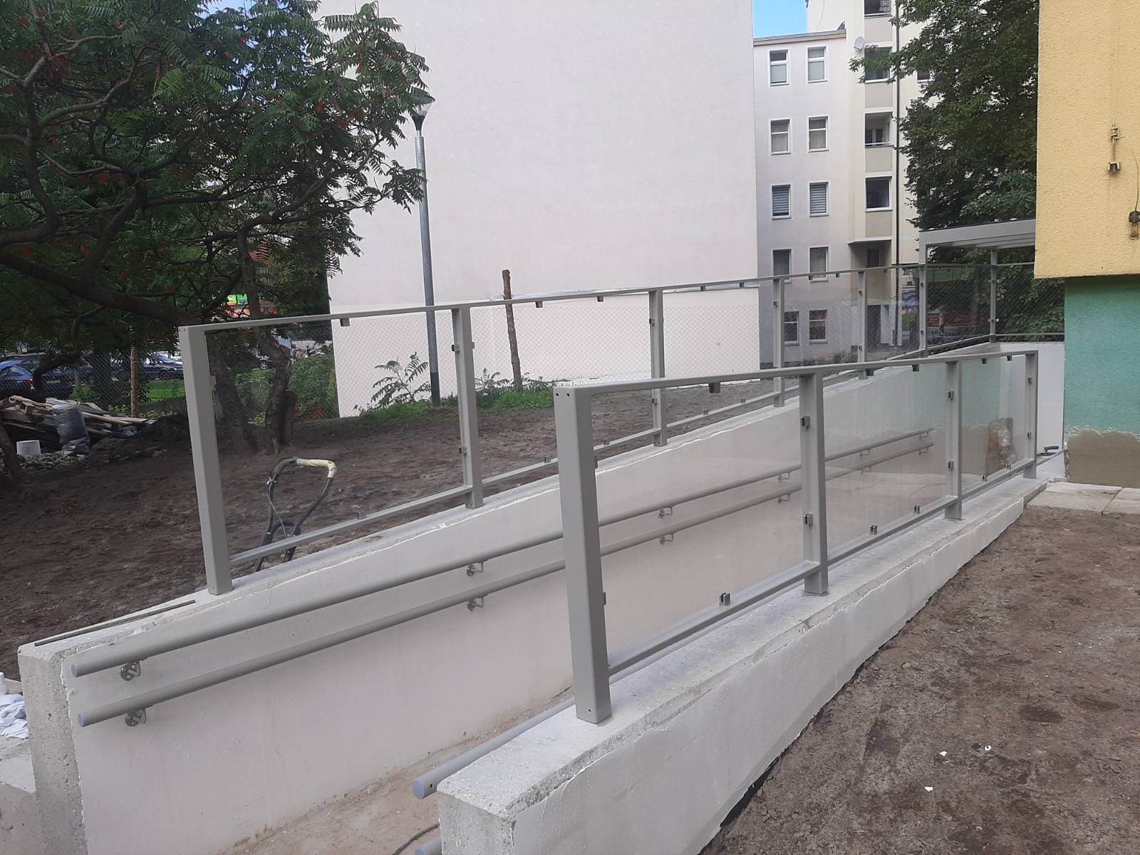 Zewnętrzna balustrada szklana z metalowymi poręczami zamontowana przy betonowym podjeździe dla wózków inwalidzkich, widok na budynek mieszkalny w tle.