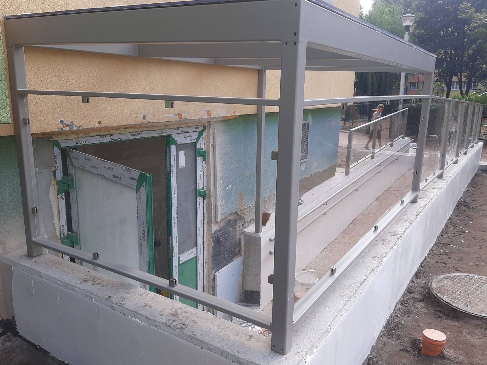 Zadaszone wejście do budynku w Szczecinie z rampą dla niepełnosprawnych, zabezpieczoną szklanymi balustradami i metalową konstrukcją.