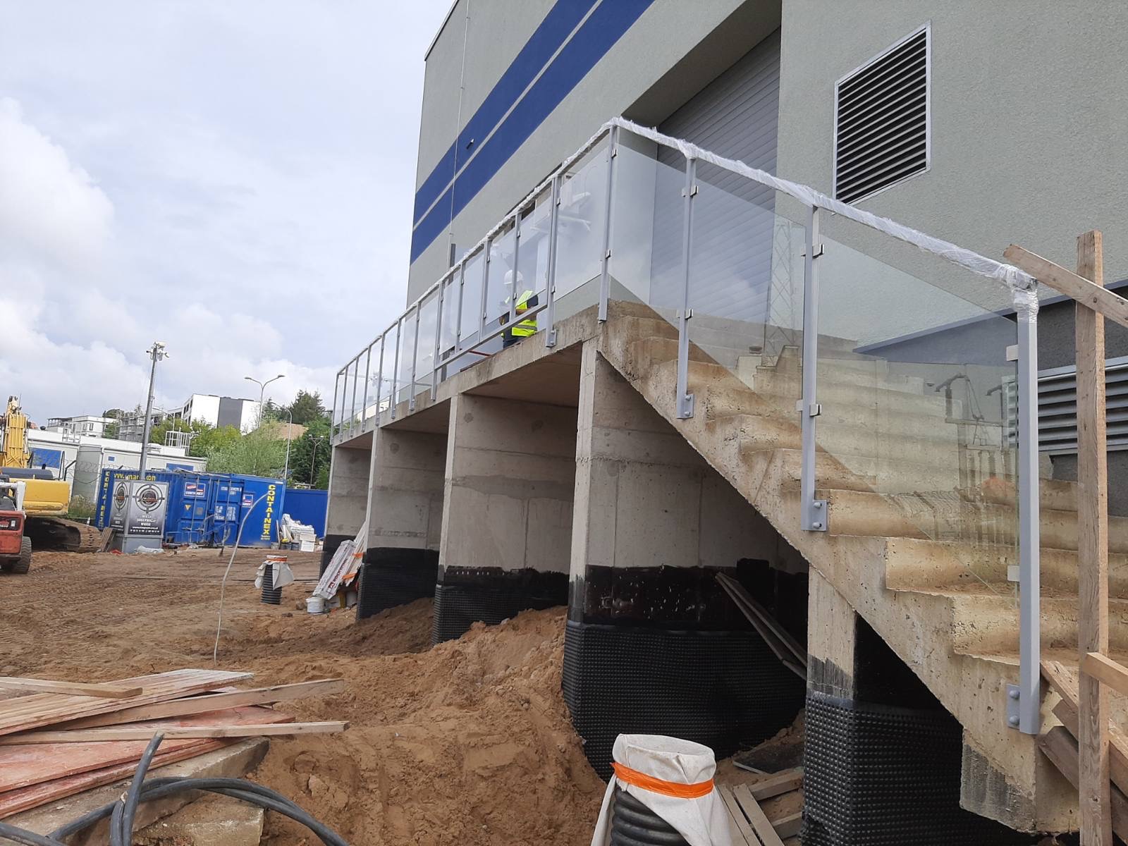 Betonowe schody zewnętrzne z balustradą szklaną w trakcie montażu, zabezpieczona folią, widoczne prace ziemne i budynek w tle.