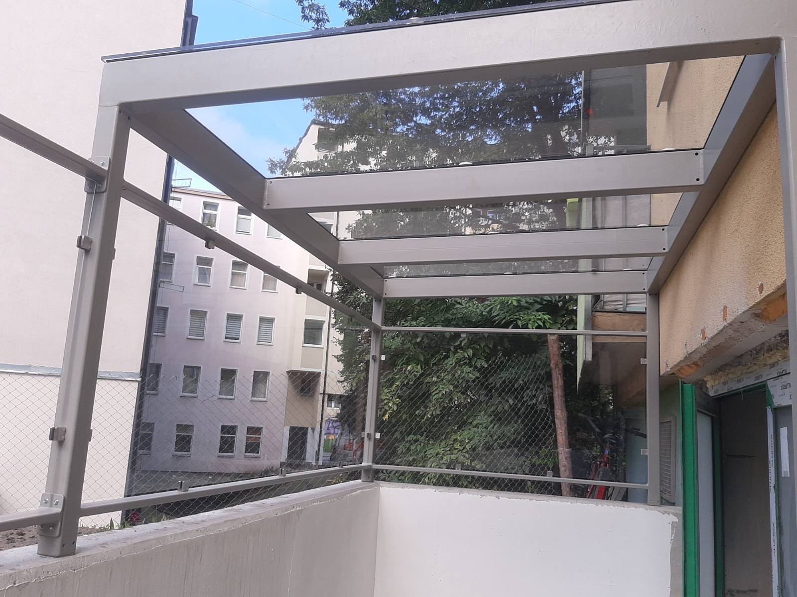 Zadaszony balkon z metalową konstrukcją i szklanym dachem, zabezpieczony siatką, widok na budynek mieszkalny i zieleń.