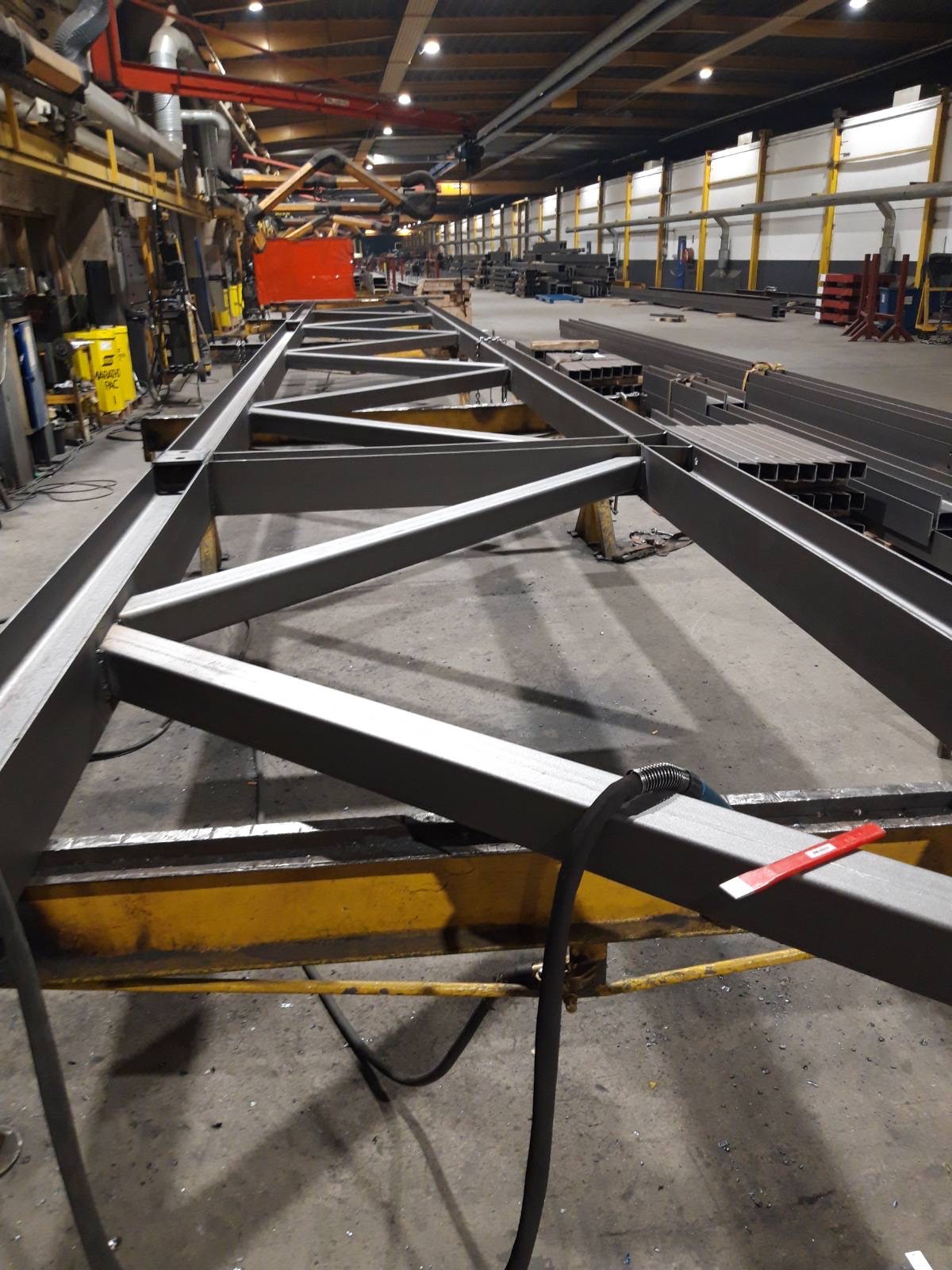 Metalowa konstrukcja kratowa, prawdopodobnie element hali, na żółtym wózku warsztatowym w hali produkcyjnej, widoczne spawarki i profile stalowe w tle.