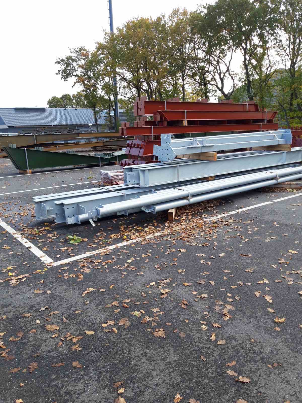 Stalowe elementy konstrukcyjne w różnych kolorach (srebrny, czerwony, zielony) ułożone na parkingu z opadłymi liśćmi, przygotowane do montażu.