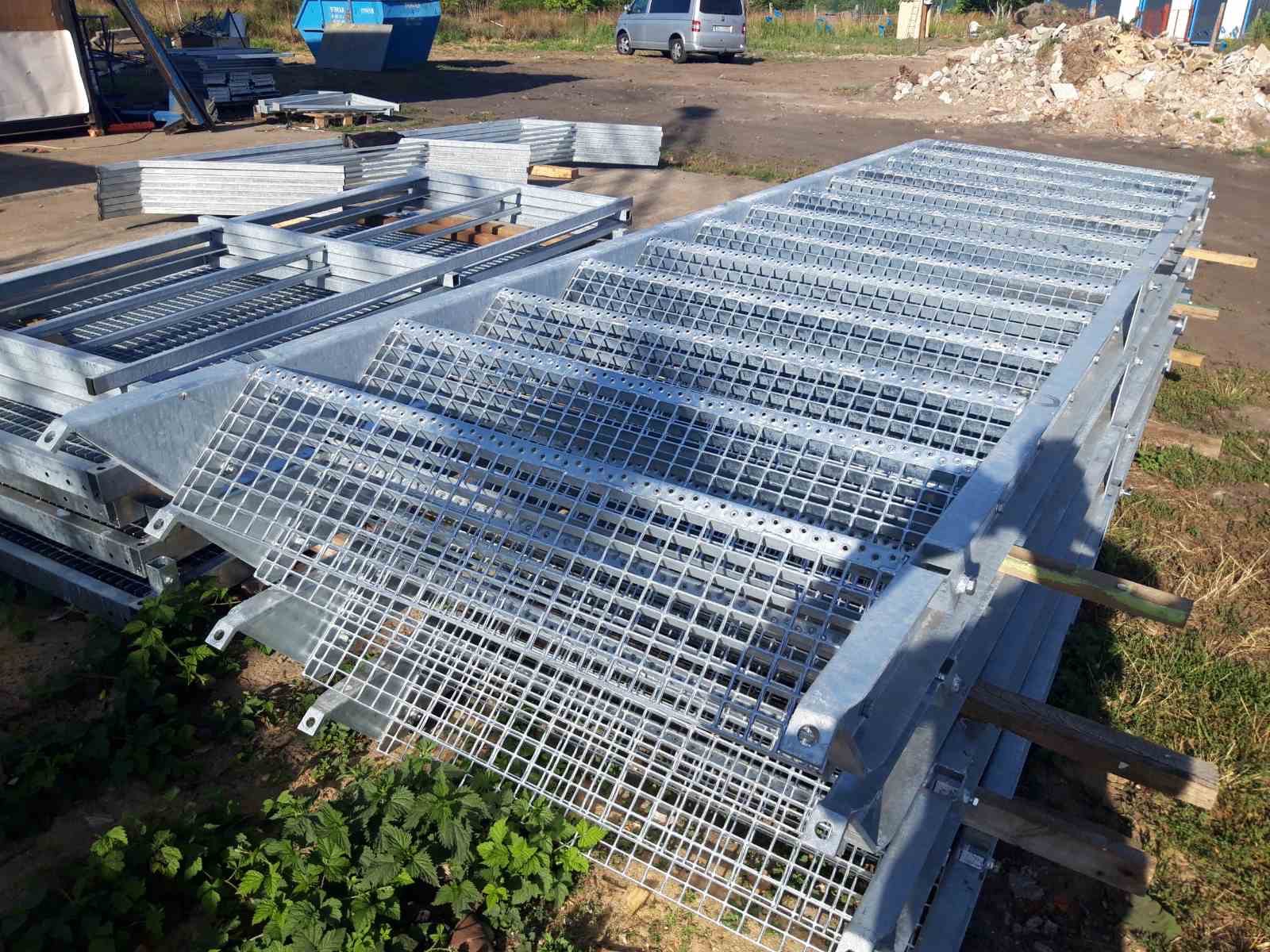 Stos ocynkowanych stopni metalowych z kratownicą, przygotowanych do montażu, na placu budowy; w tle widoczny kontener i samochód dostawczy.