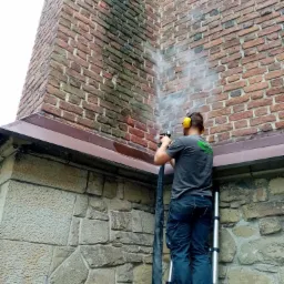 Pracownik w słuchawkach ochronnych czyści ceglaną elewację budynku z kamienną podmurówką za pomocą myjki ciśnieniowej, widoczny efekt czyszczenia.