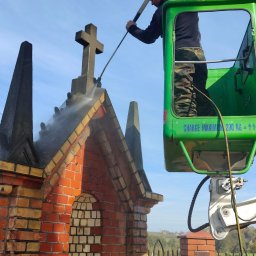 Cps technology - Pracownik w podnośniku koszowym czyści ciśnieniowo zabytkową, ceglaną kapliczkę z krzyżem na dachu.