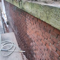 Cps technology - Częściowo oczyszczona ceglana elewacja, widoczne różnice w kolorze i zabrudzeniu, wąż z dyszą leżący na betonowej powierzchni, góra muru porośnięta roślinnością.