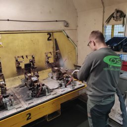 Cps technology - Pracownik w okularach czyści sprężonym powietrzem metalowe elementy na żółtym stole warsztatowym z numerem 2, w tle żółta ściana i logo firmy na bluzie pracownika.