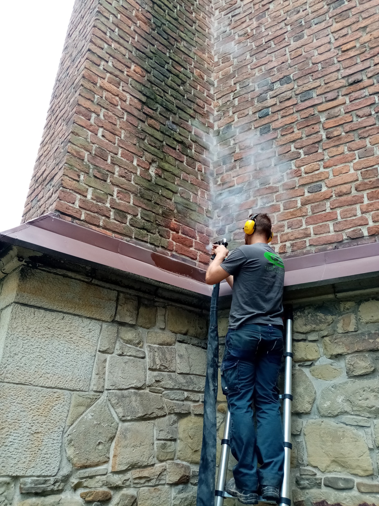 Pracownik w słuchawkach ochronnych czyści ceglaną elewację budynku z kamienną podmurówką za pomocą myjki ciśnieniowej, widoczny efekt czyszczenia.