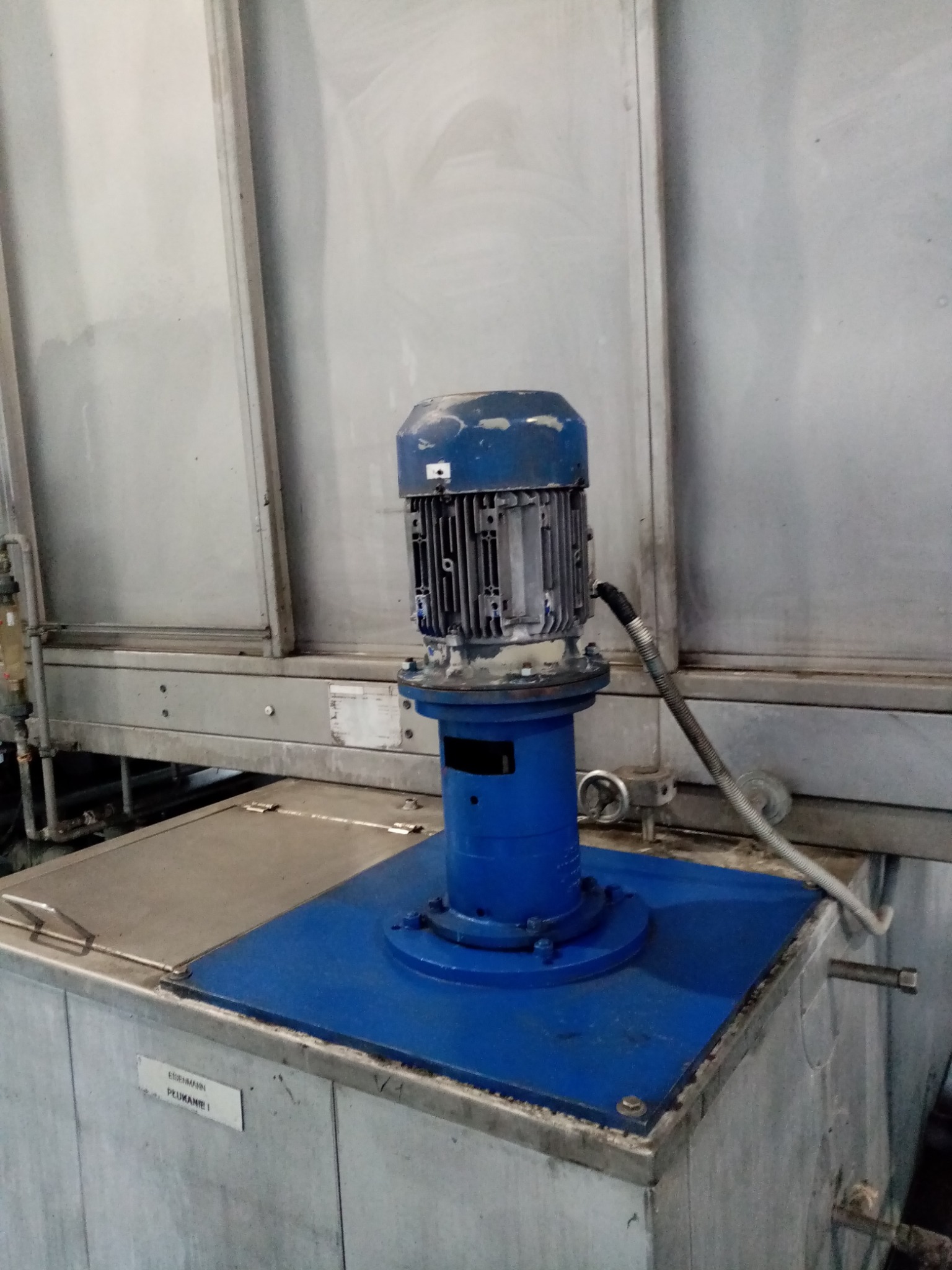 Niebieski silnik elektryczny na metalowej podstawie, element instalacji przemysłowej w hali, widoczne rury i metalowe ściany.