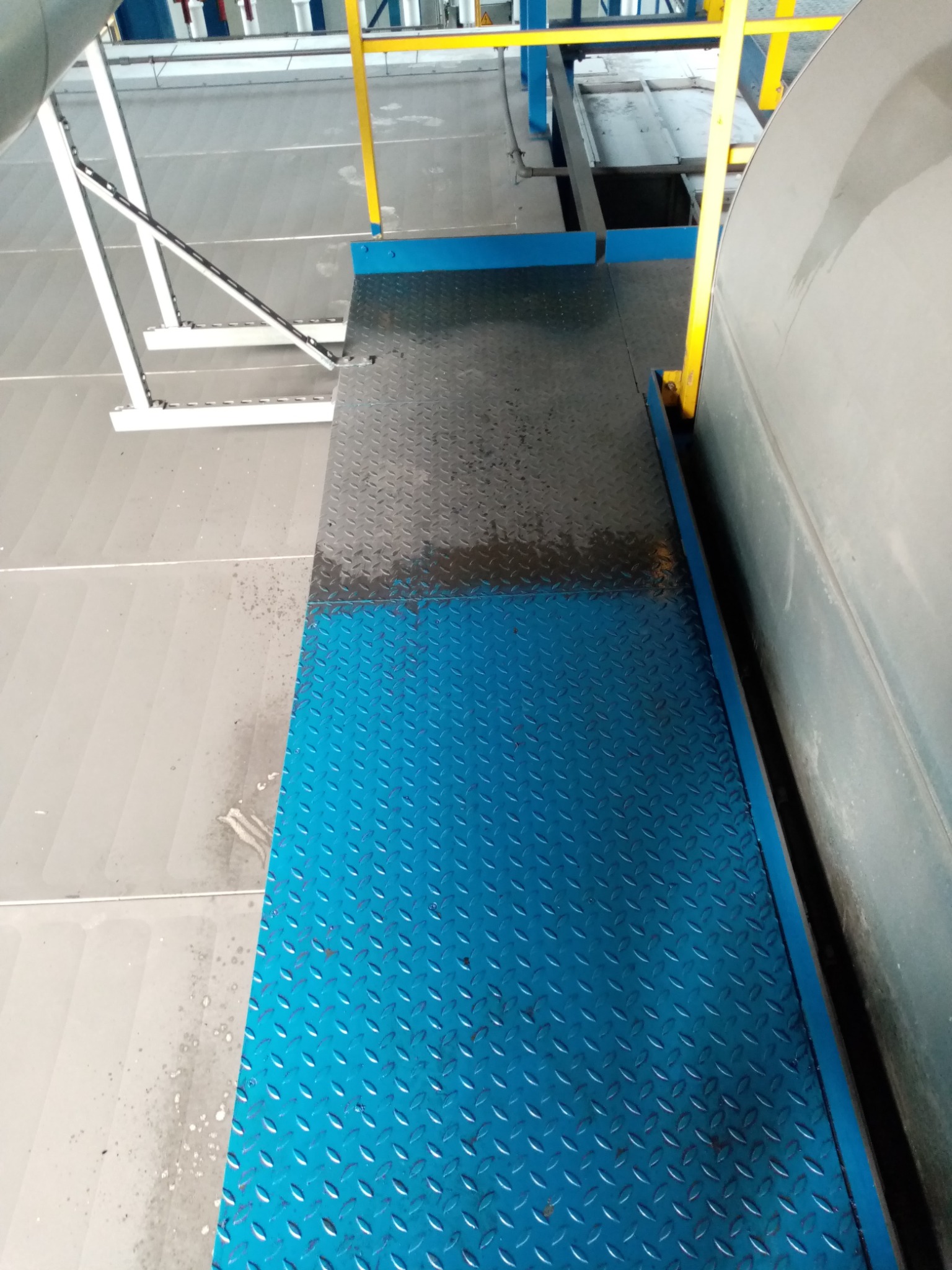 Niebieska, metalowa platforma z antypoślizgową fakturą, przylegająca do szarego, cylindrycznego zbiornika w hali przemysłowej, z widocznymi elementami konstrukcyjnymi w kolorach żółtym i białym.
