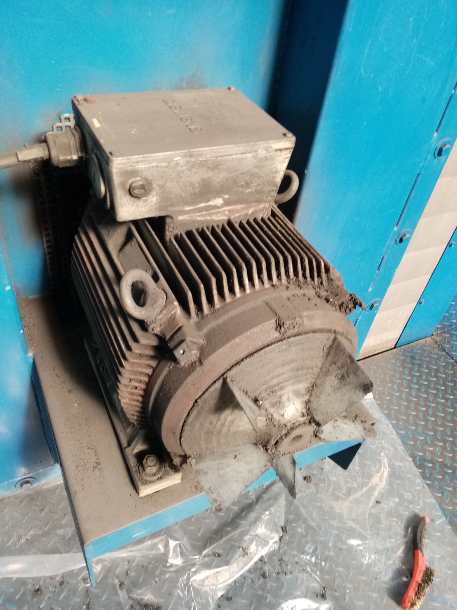 Zabrudzony silnik elektryczny z wentylatorem, osadzony na metalowej podstawie, widoczny kabel zasilający i szczotka do czyszczenia w tle.