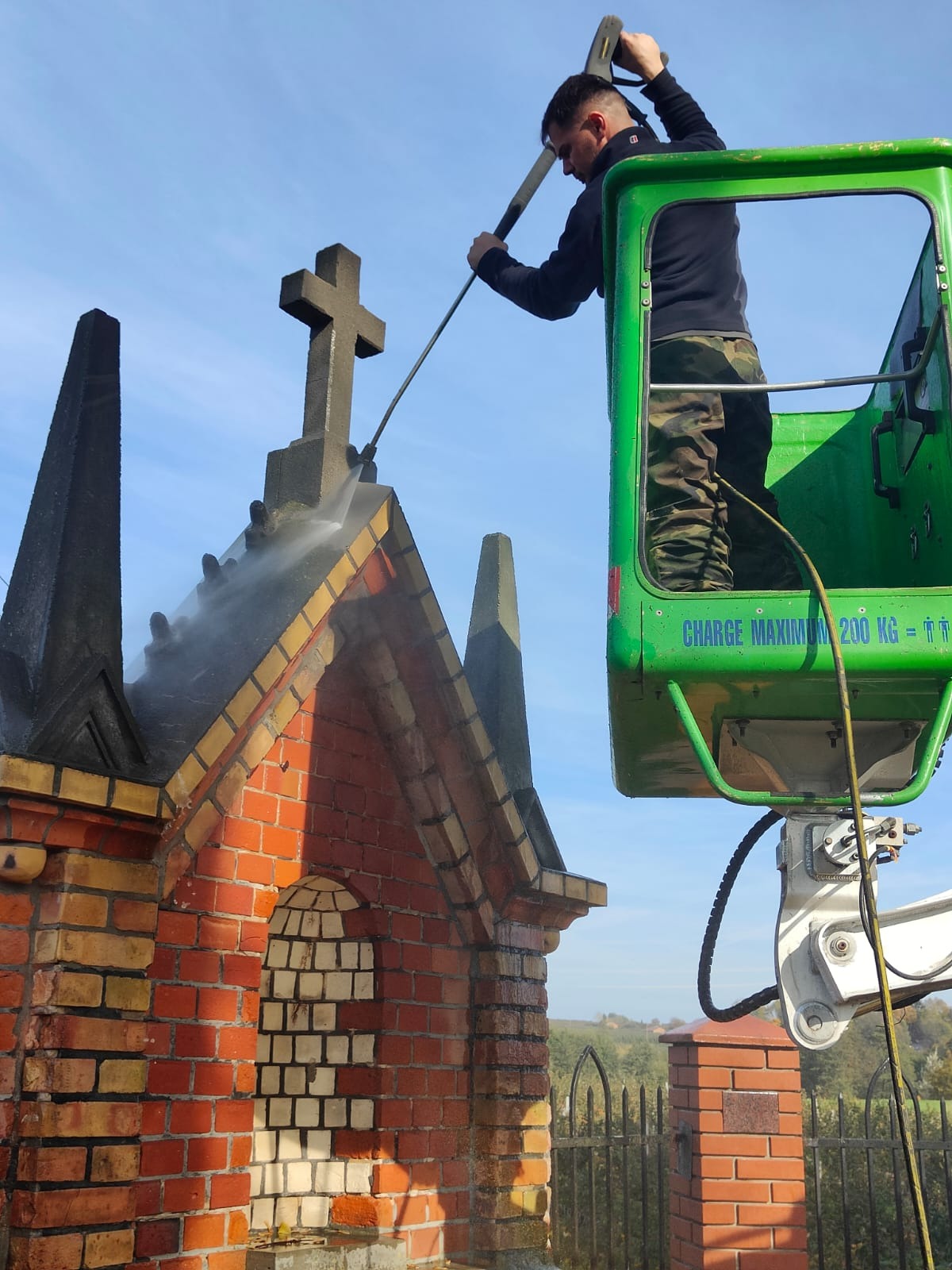 Pracownik w podnośniku koszowym czyści ciśnieniowo zabytkową, ceglaną kapliczkę z krzyżem na dachu.