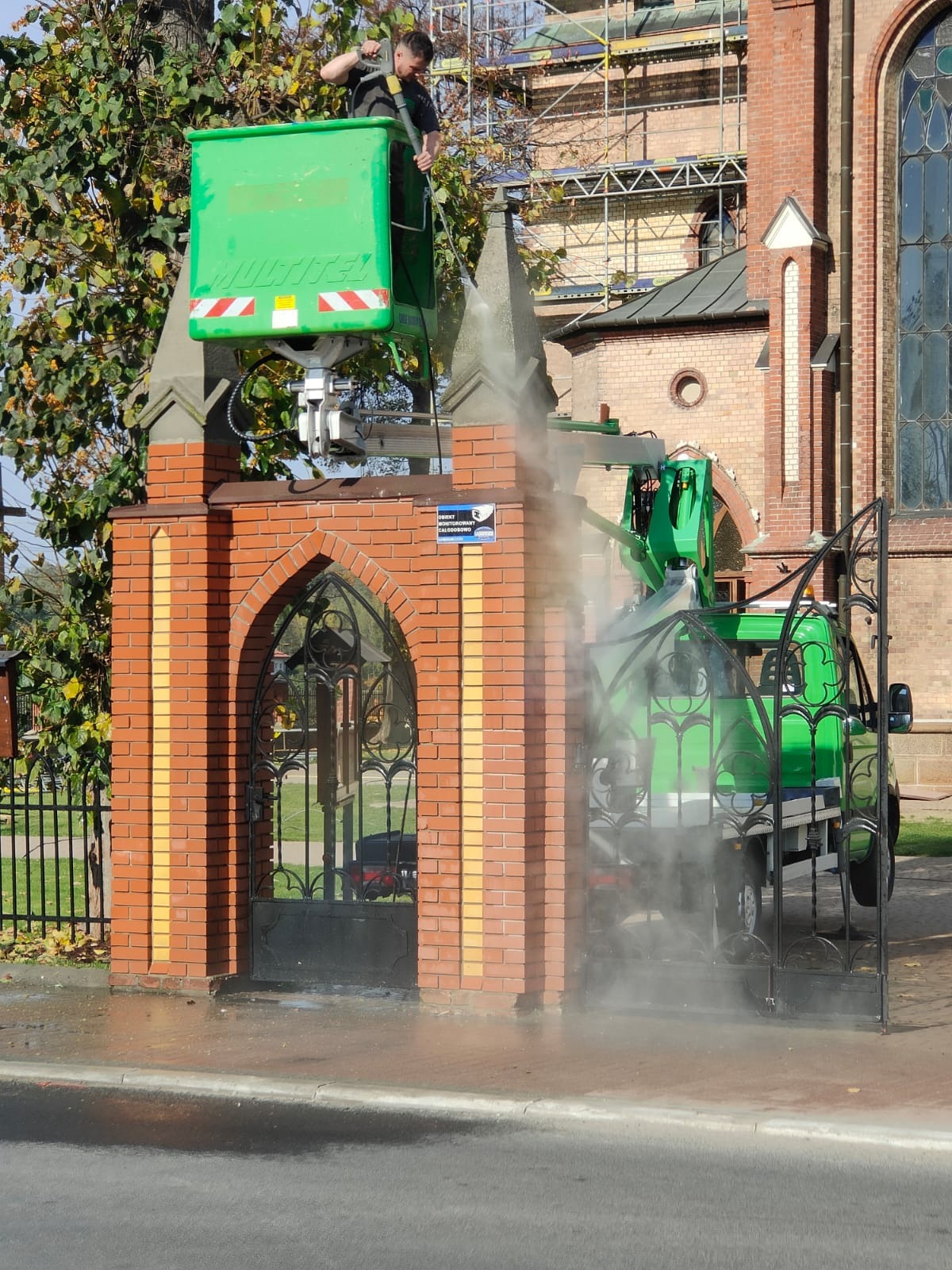 Pracownik w zielonym podnośniku koszowym myje ciśnieniowo ceglany mur z ozdobną bramą wjazdową, na tle kościoła w rusztowaniach.