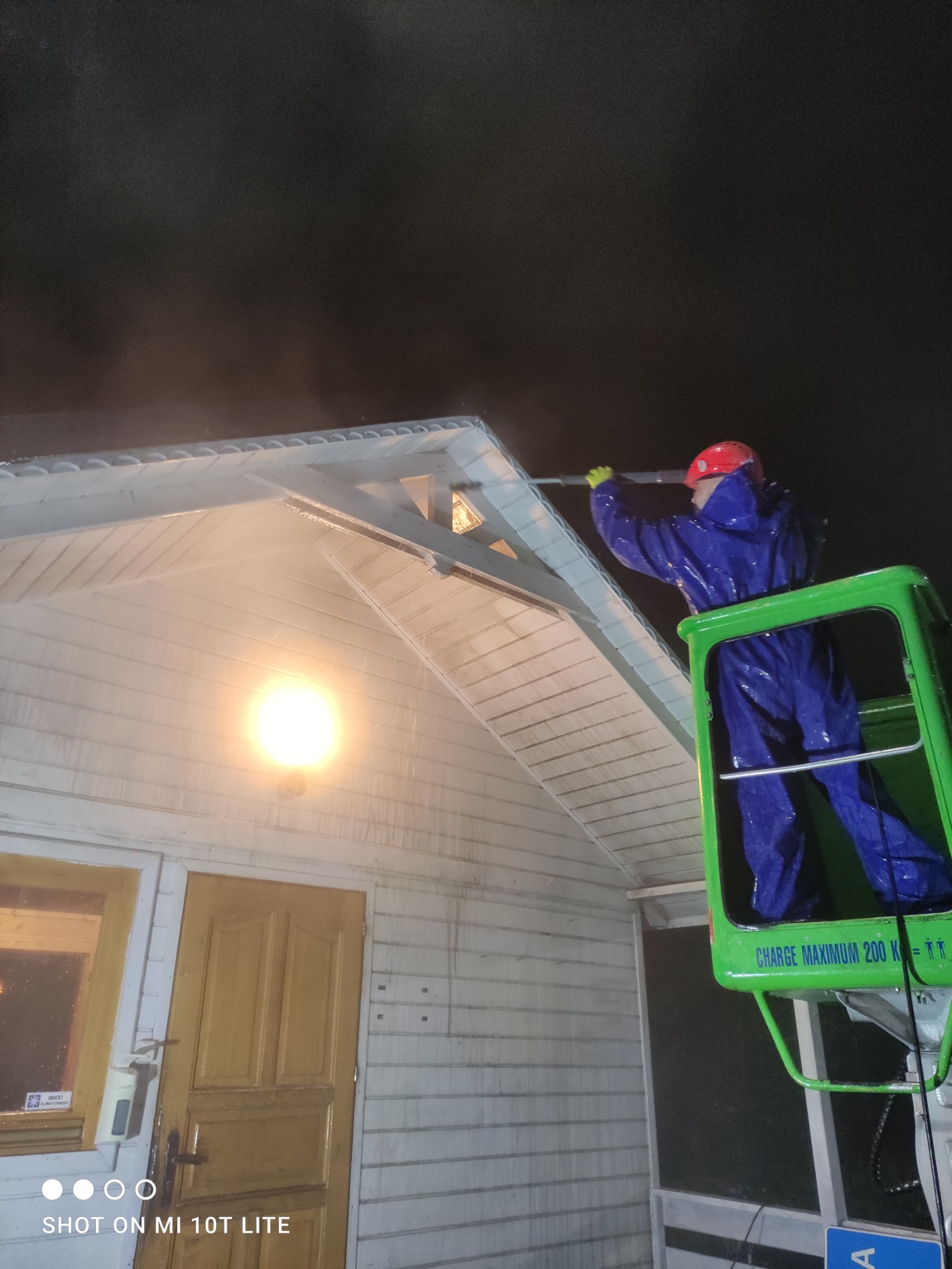 Pracownik w niebieskim kombinezonie i czerwonym kasku, stojący w podnośniku koszowym, myje elewację domu pod silnym strumieniem wody pod ciśnieniem, widoczne zabrudzenia na elewacji przed umyciem...