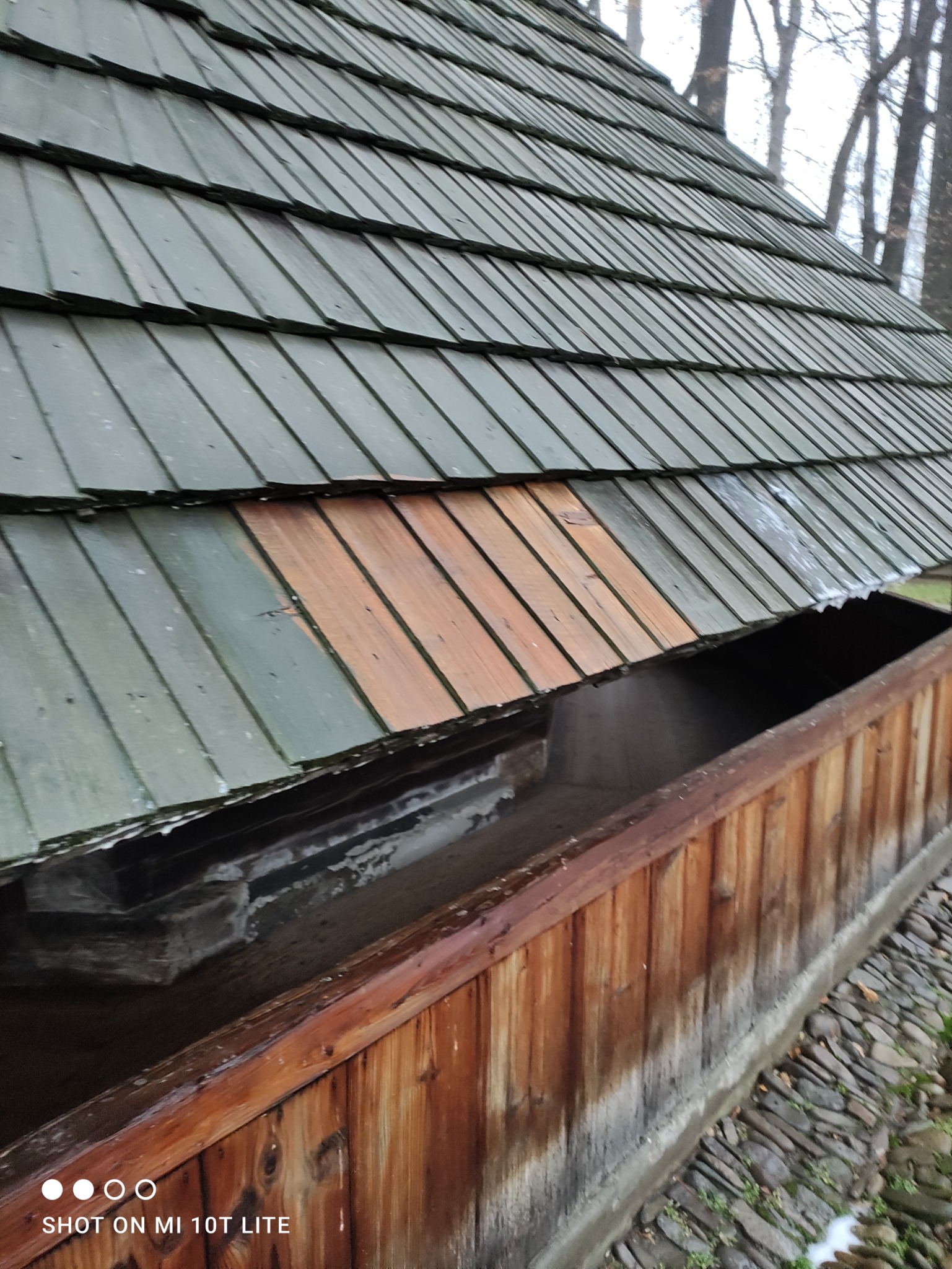 Fragment drewnianego dachu z rynną, częściowo pomalowanego na zielono, z fragmentem niezabezpieczonego drewna. Widoczne ślady wilgoci i zabrudzenia.