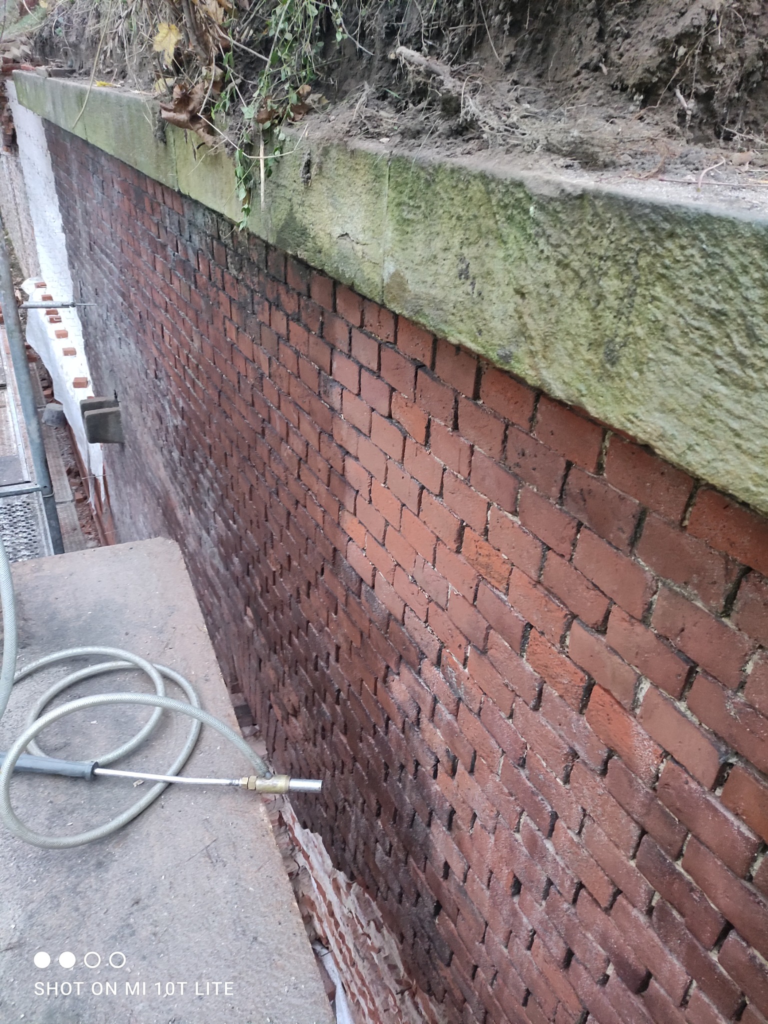 Częściowo oczyszczona ceglana elewacja, widoczne różnice w kolorze i zabrudzeniu, wąż z dyszą leżący na betonowej powierzchni, góra muru porośnięta roślinnością.