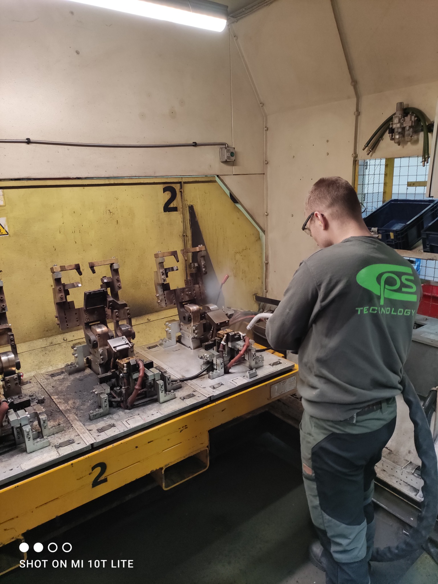 Pracownik w okularach czyści sprężonym powietrzem metalowe elementy na żółtym stole warsztatowym z numerem 2, w tle żółta ściana i logo firmy na bluzie pracownika.