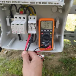 Pomiar napięcia multimetrem NEO na zabezpieczeniach przeciwprzepięciowych w skrzynce elektrycznej instalacji fotowoltaicznej; widoczne przewody, listwa zaciskowa i elementy instalacji na zewnątrz.