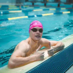 Uśmiechnięty mężczyzna w r&oacute;żowym czepku i okularach pływackich, oparty o brzeg basenu, pokazuje kciuki w g&oacute;rę