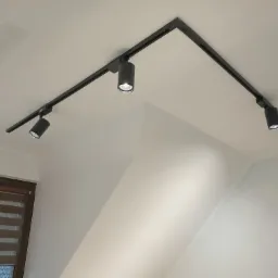 Czarna szyna oświetleniowa z czterema reflektorami punktowymi zamontowana na białym, skośnym suficie poddasza, widoczna z okna z żaluzjami dzień i noc.