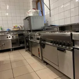 Profesjonalna kuchnia z nierdzewnymi urządzeniami, widoczne płyty grzewcze, piekarnik, zlew i okap, białe kafelki na ścianach i podłodze, kosz z praniem na piekarniku.