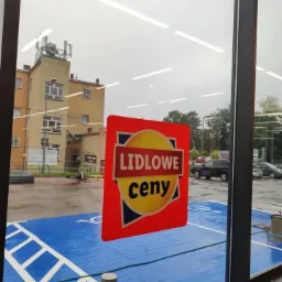 Czerwona naklejka z napisem 'LIDLOWE ceny' na szybie sklepu, widok na mokry parking z niebieską kostką brukową i budynek w tle.