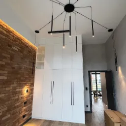 Nowoczesne oświetlenie sufitowe z wieloma czarnymi lampami punktowymi w przestronnym wnętrzu z ceglaną ścianą i białą szafą.