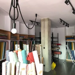 Wnętrze sklepu z tkaninami, widoczne rolki materiałów na półkach i w ekspozycji, oświetlenie szynowe i lampa wisząca na grubym kablu.