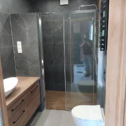 Nowoczesna łazienka z kabiną prysznicową typu walk-in, umywalką nablatową na drewnianej szafce, wiszącą toaletą i czarnym grzejnikiem drabinkowym; widoczne szare płytki imitujące kamień...