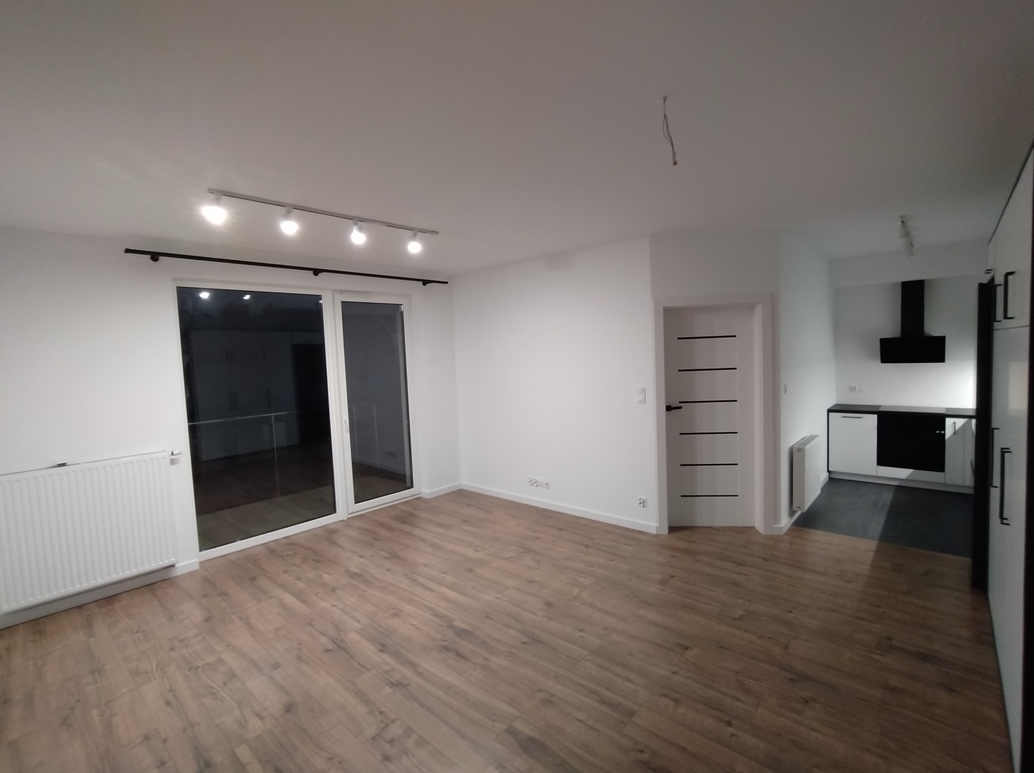 Jasne, przestronne wnętrze po remoncie: parkiet w odcieniach brązu, białe ściany, grzejnik, okno balkonowe z czarną ramą, białe drzwi z poziomymi frezami, widok na kuchnię z czarnym okapem.