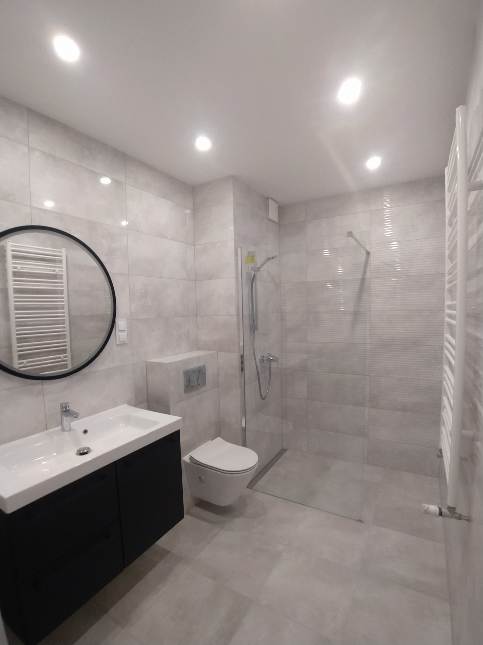 Nowoczesna łazienka z szarymi płytkami, czarną szafką pod umywalką z okrągłym lustrem i kabiną prysznicową typu walk-in z szklaną ścianką.