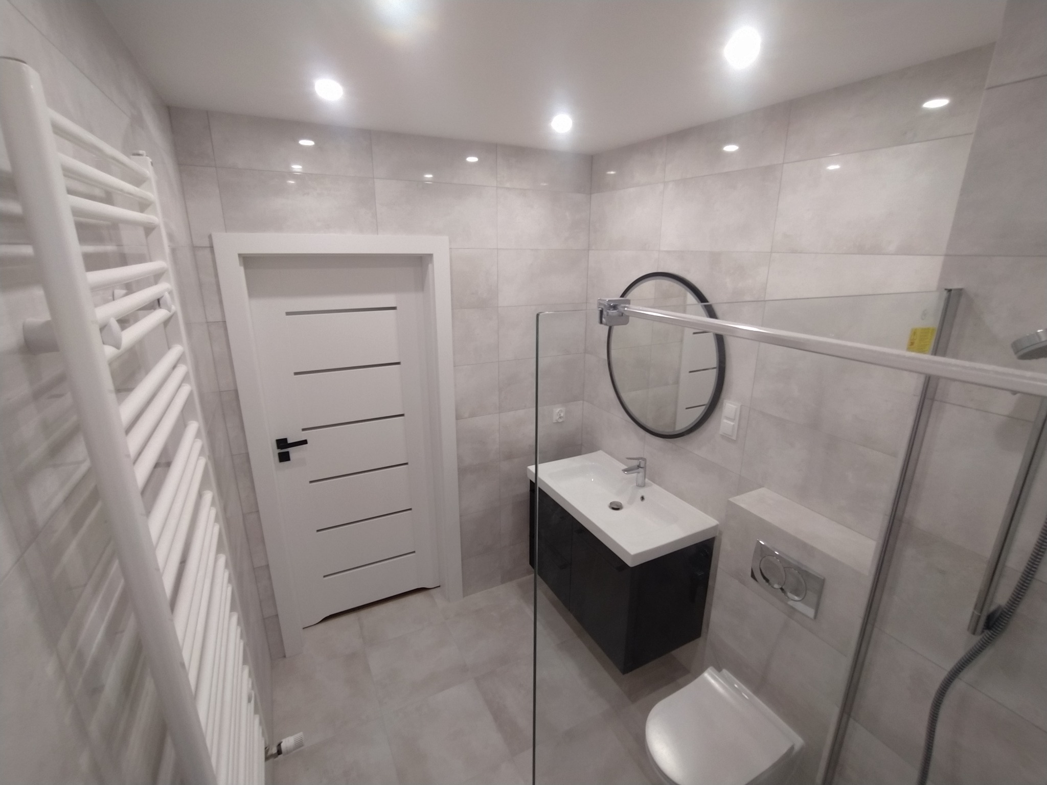 Nowoczesna łazienka z płytkami imitującymi kamień, wiszącą szafką z umywalką, okrągłym lustrem w czarnej ramie, białym grzejnikiem drabinkowym i kabiną prysznicową ze szklaną ścianką.