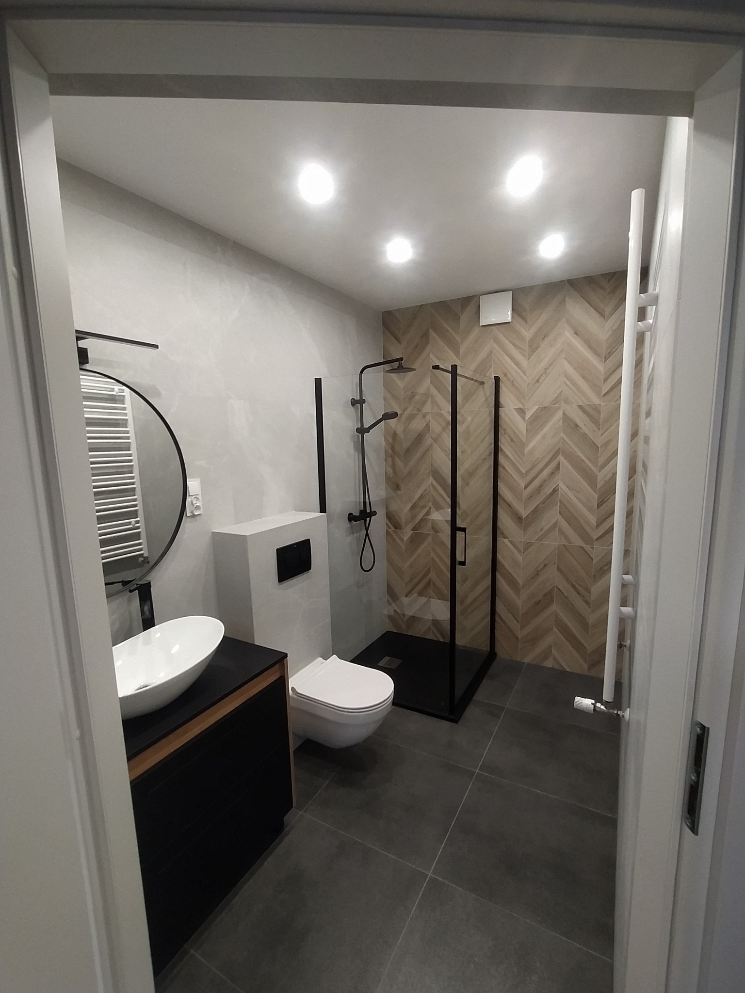 Nowoczesna łazienka z kabiną prysznicową typu walk-in z czarnymi ramami, umywalką nablatową na czarnej szafce z drewnianymi akcentami, okrągłym lustrem z czarną ramą i grzejnikiem drabinkowym w tle...