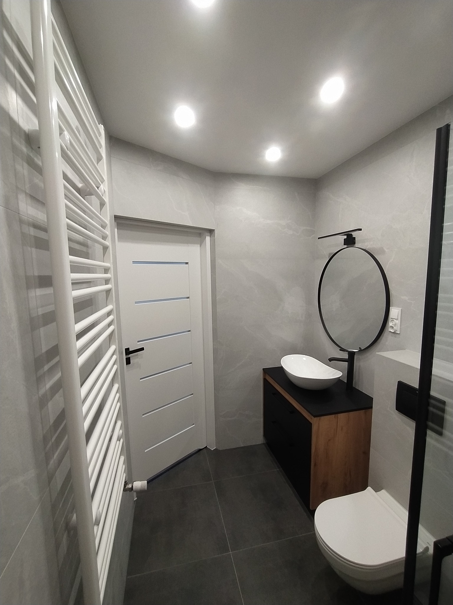 Nowoczesna łazienka w szarościach z białą ceramiką, owalną umywalką nablatową i okrągłym lustrem z czarną ramą, grzejnik drabinkowy po lewej stronie, drzwi z poziomymi wgłębieniami.