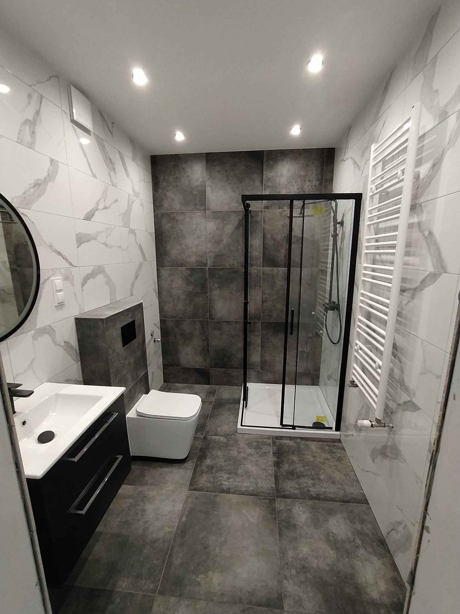 Nowoczesna łazienka z kabiną prysznicową w czarnej ramie, umywalką z szafką w kolorze czarnym, toaletą w zabudowie i grzejnikiem drabinkowym na tle płytek imitujących marmur.