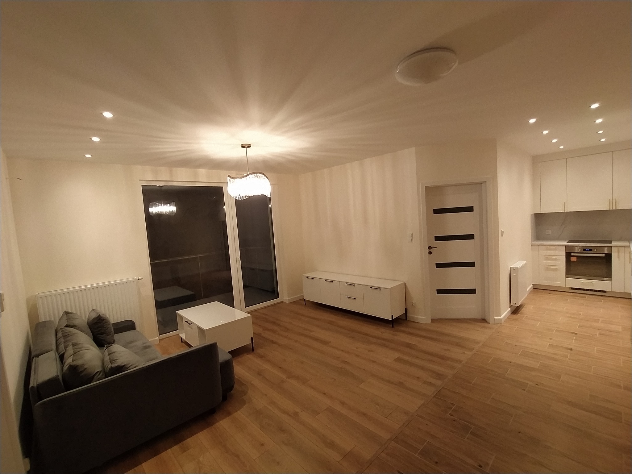 Jasne, przestronne wnętrze mieszkania po remoncie z drewnianą podłogą, szarą sofą, białymi meblami i oświetleniem punktowym oraz wiszącą lampą.