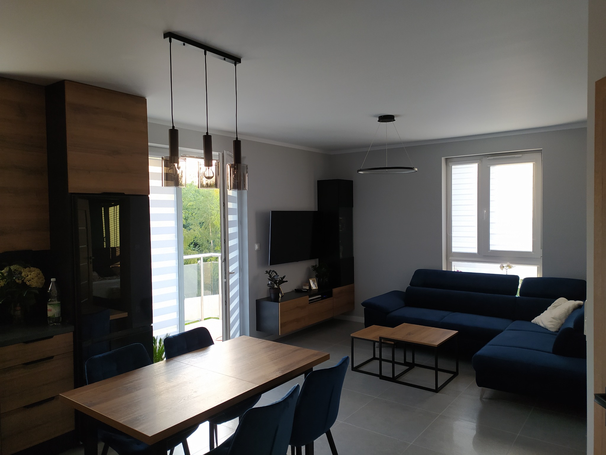 Jasne wnętrze salonu z jadalnią, szare ściany, drewniany stół z welurowymi krzesłami, narożna sofa w odcieniach granatu, podwieszane lampy nad stołem i okrągła lampa sufitowa w części wypoczynkowej...