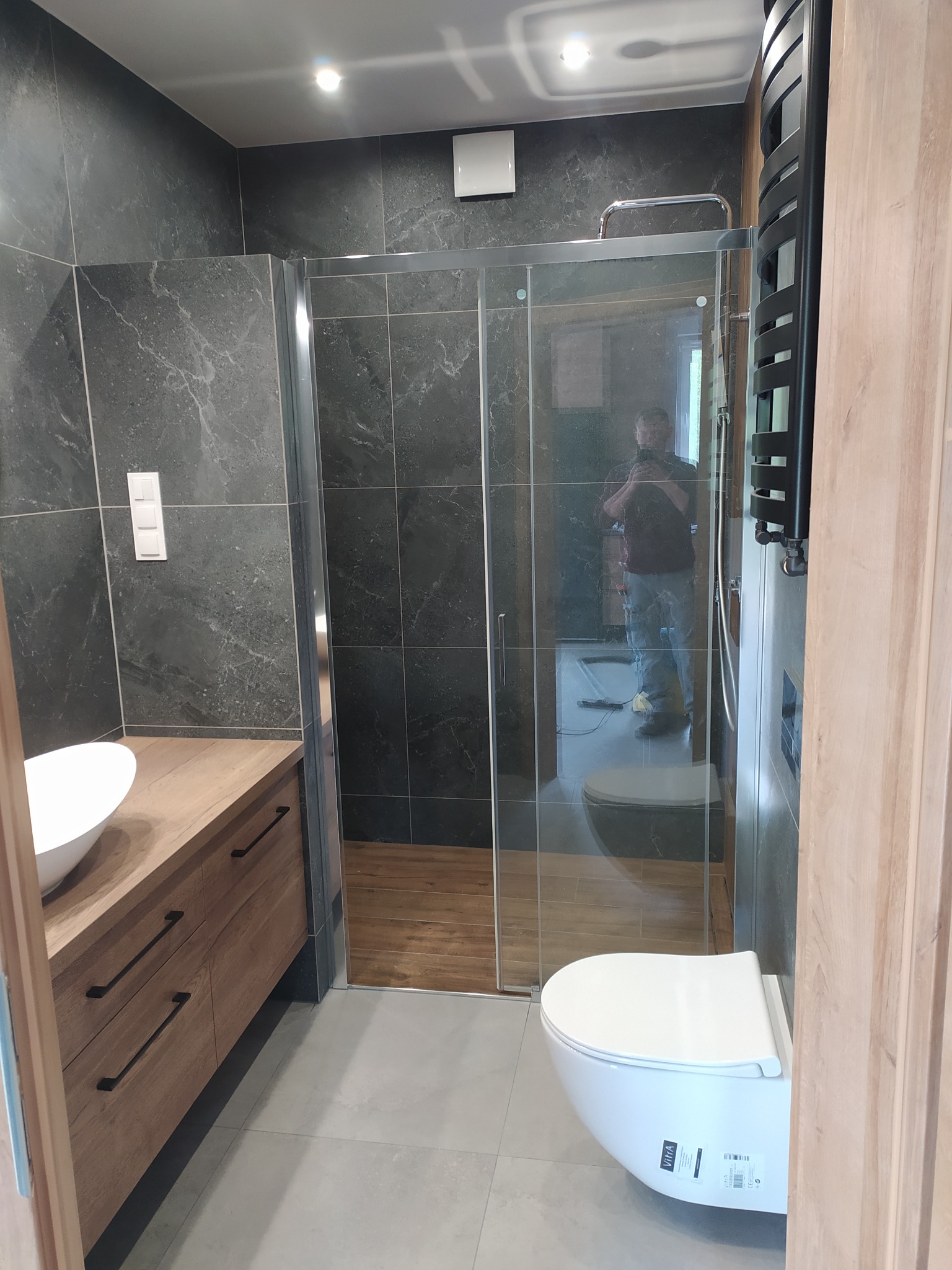 Nowoczesna łazienka z kabiną prysznicową typu walk-in, umywalką nablatową na drewnianej szafce, wiszącą toaletą i czarnym grzejnikiem drabinkowym; widoczne szare płytki imitujące kamień...