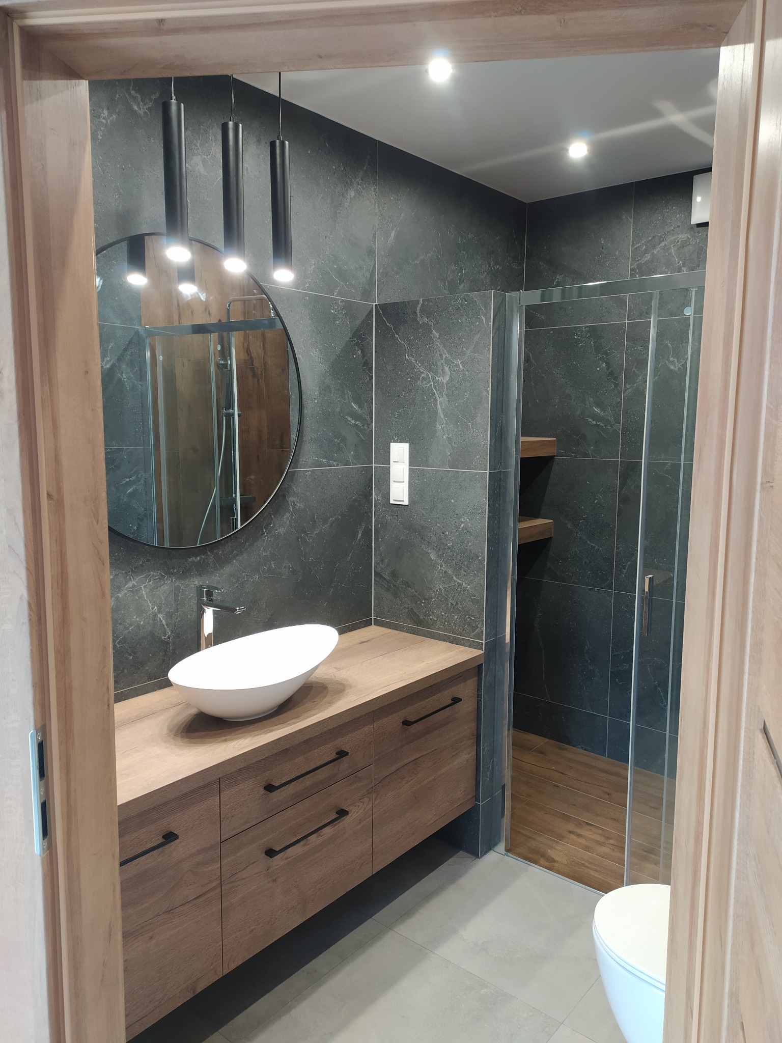 Nowoczesna łazienka z ciemnymi płytkami imitującymi marmur, drewnianą szafką pod umywalką z minimalistycznymi uchwytami, okrągłym lustrem i kabiną prysznicową z drewnianymi półkami.
