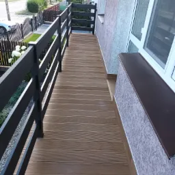 Długi, wąski balkon z brązową, kompozytową podłogą imitującą drewno, oraz ciemnoszarą, nowoczesną balustradą. Widok na ulicę z zabudową jednorodzinną i ogrodami.