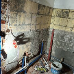 T home Tomasz Bokuniewicz - Łazienka w trakcie remontu: odkryte instalacje wodne, ściany z częściowo usuniętymi płytkami, poziomica oparta o ścianę, wiadro z silikonem, narzędzia leżące na podłodze.