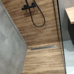 Nowoczesny prysznic typu walk-in z drewnopodobnymi płytkami na podłodze i czarną armaturą, widoczny odpływ liniowy i szklana ścianka kabiny.