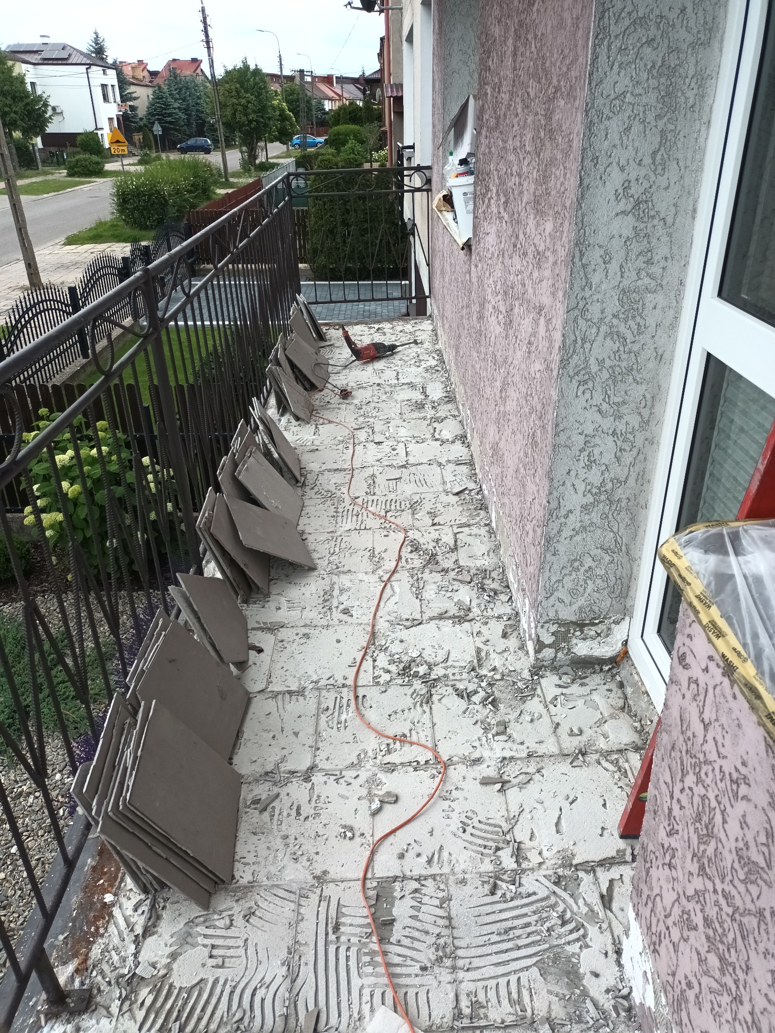 Balkon w trakcie demontażu starej terakoty, widoczne stosy płytek, resztki kleju, szlifierka kątowa i przewód zasilający, balustrada balkonowa z widokiem na ulicę.